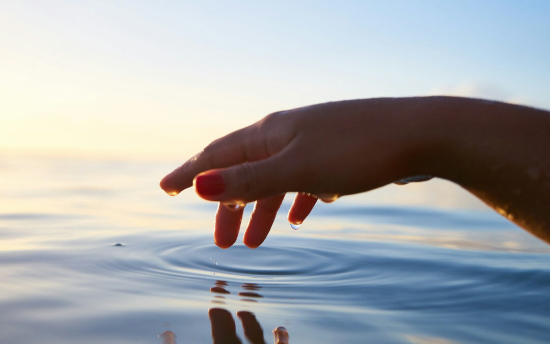 Una mano sfiora uno specchio d'acqua con un dito, creando un cerchio concentrico.